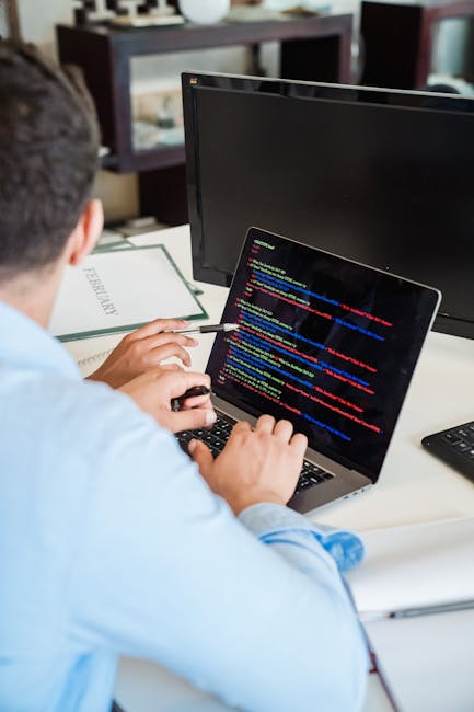 A person seated at a desk in an office environment, working on a laptop displaying lines of colorful computer code. The desk has a black computer monitor, a closed notebook labeled 'PERFORMANCE,' and