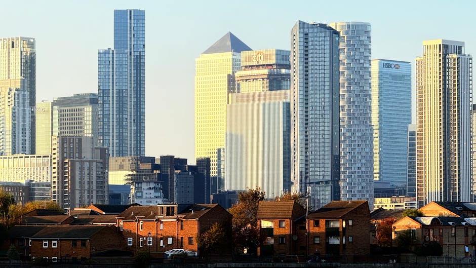 A cityscape featuring a row of red-brick residential houses in the foreground, with a modern commercial skyscraper district in the background. The tall glass and steel buildings reflect sunlight, show